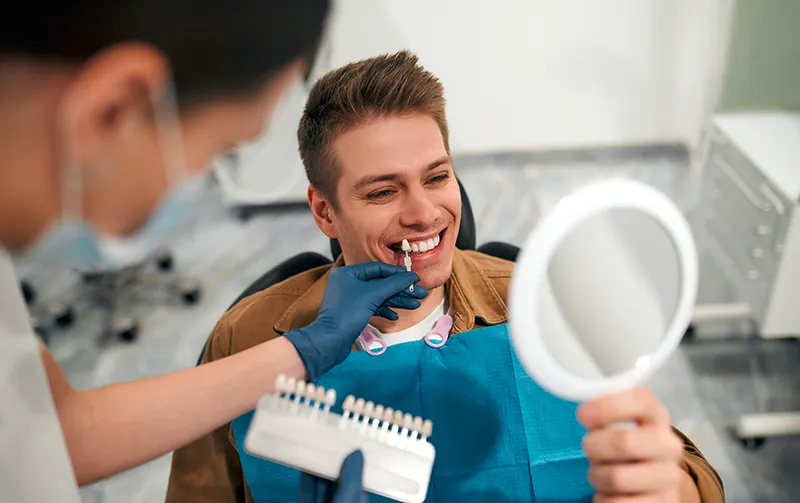 Young man checking his smile in a mirror while the dentist matches a veneer shade guide to his teeth